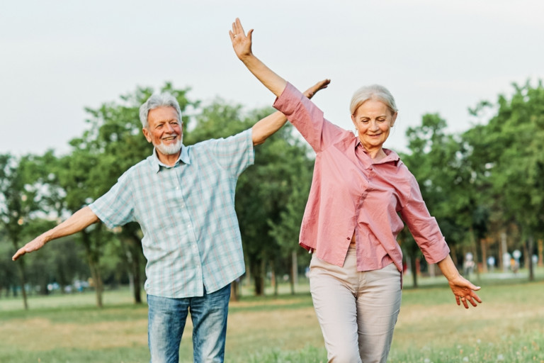 Happy senior couple goofing around with outstretched arms trying to walk an imaginary balance beam outside.