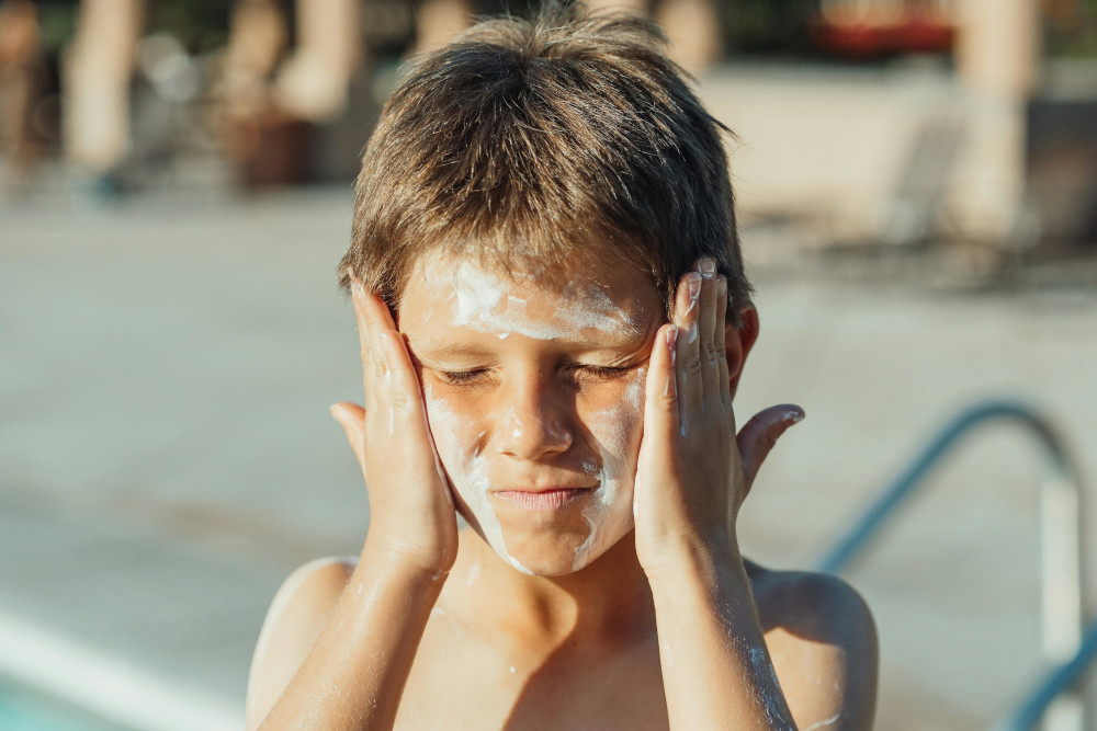 Boy applying sunscreen to his face beside a public swimming pool.