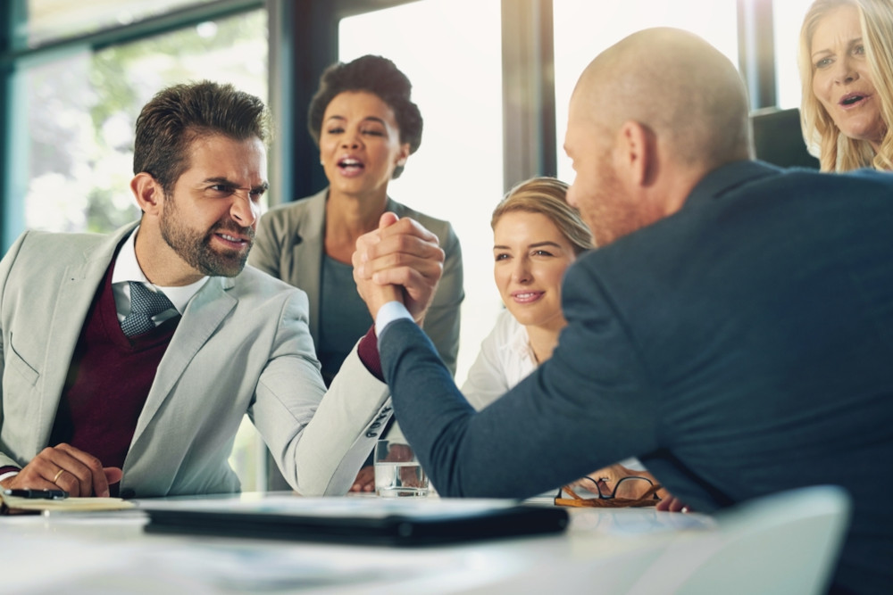 Two men in suits arm wrestling in an office while co-workers cheer them on.