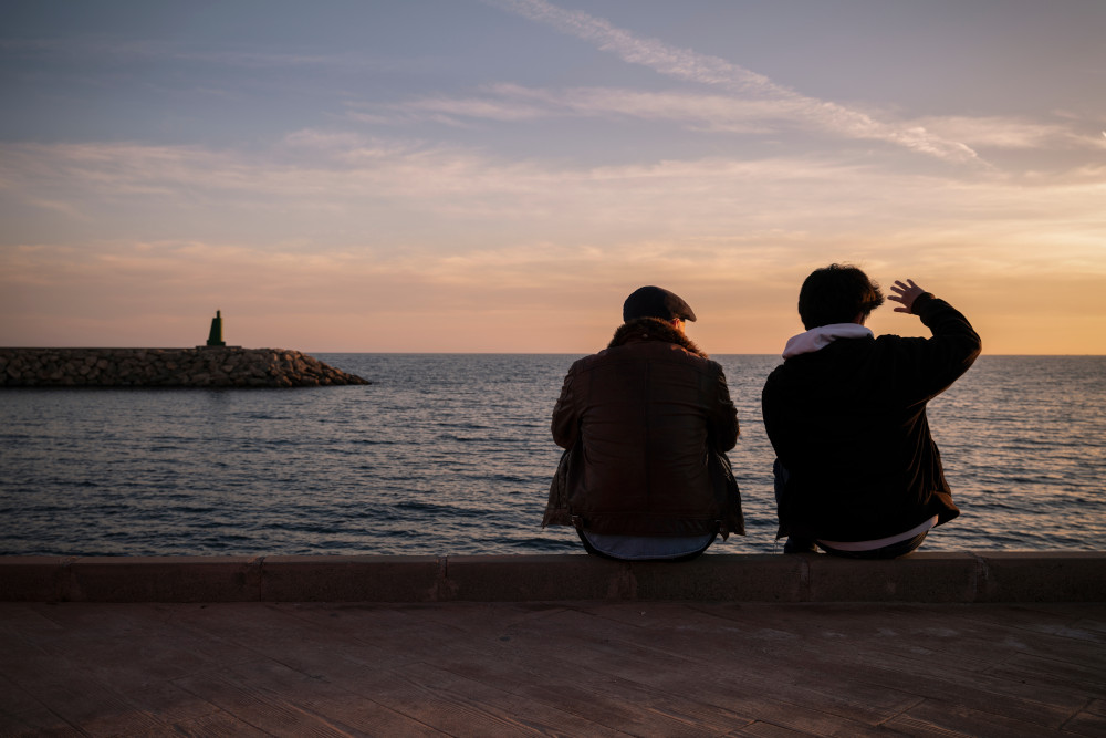 Father and son sitting together at dusk watching the sunset by the ocean.