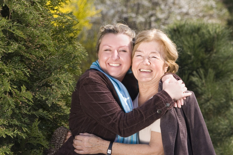 Two female family members smiling for the camera as they hug for a 'generations' photo in front of pine trees.