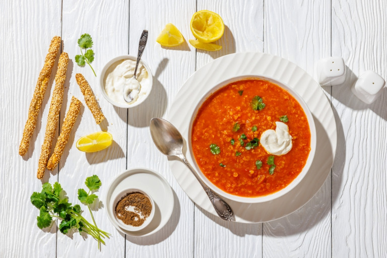 Red lentil soup in a bowl next to bread sticks, fresh cilantro, sour cream, and lemon wedges. 