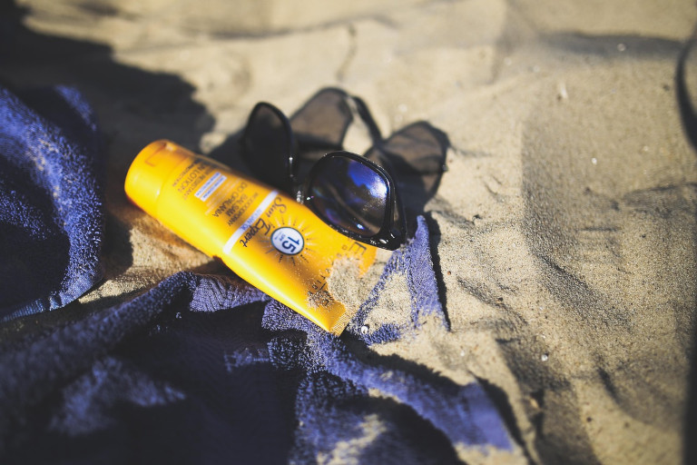 Bottle of sunscreen laying on a beach towel by a pair of sunglasses on the sand.