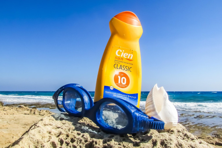 Sunscreen, swimming goggles and a seashell on a pile of sand at the beach with the ocean and skyline in the background.