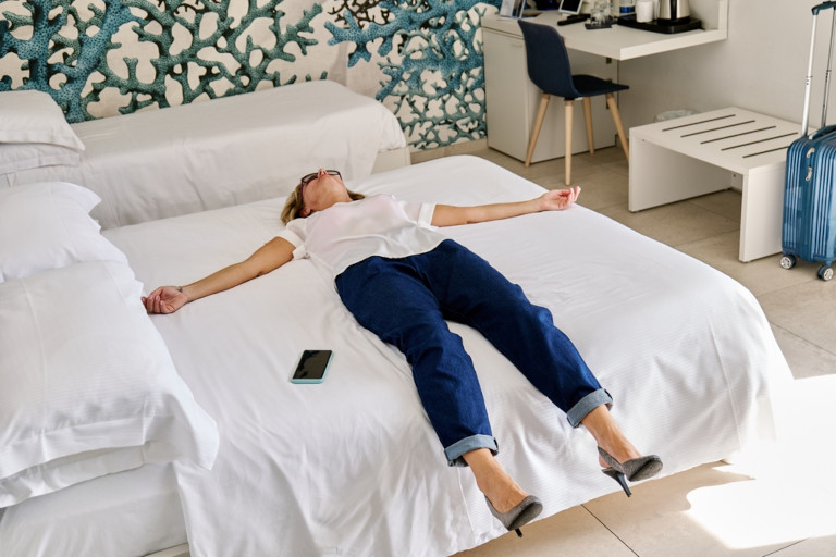 Tired female tourist sprawled out on the bed in a hotel room still wearing sunglasses and shoes.