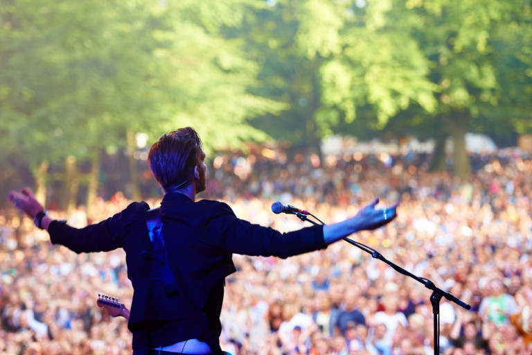 Lead singer on stage with a crowd stretched out in front of him during a daytime outdoor concert.