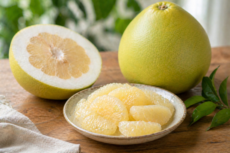 A bowl with pomelo segments sits on a table beside a whole pomelo and a pomelo that has been cut in half.