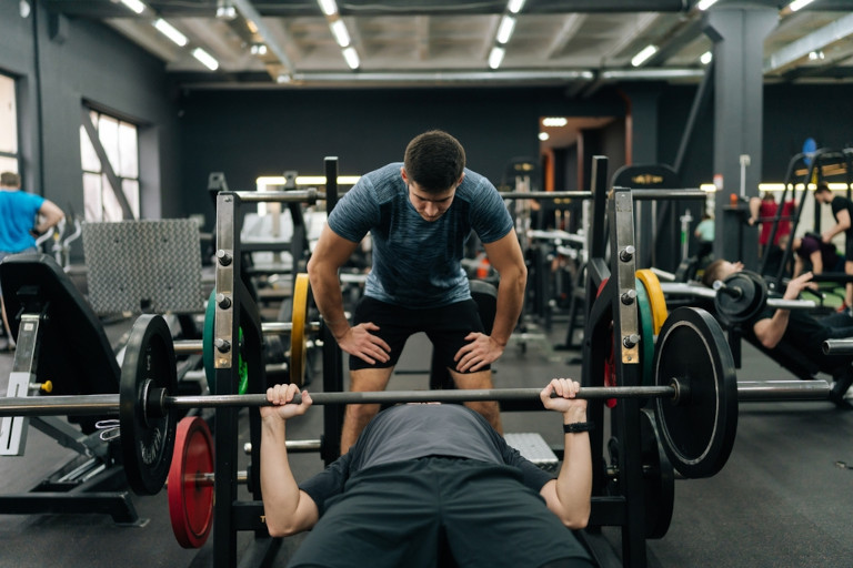 Spotter standing over a man while he bench presses 135 lbs in the gym.