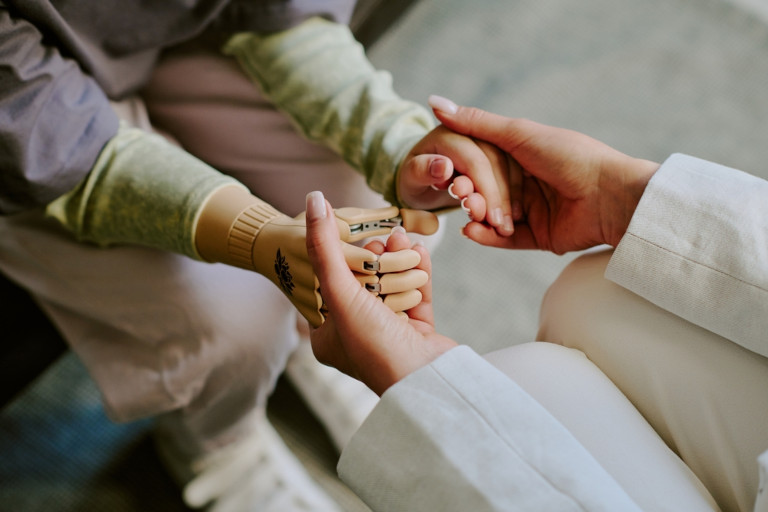 Caregiver sitting with a patient and holding both their own biological hand and their prosthetic hand.