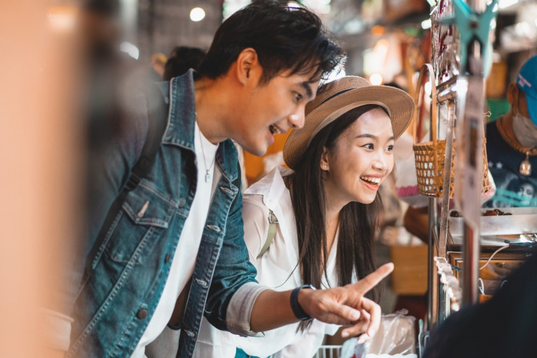 Two Asian shoppers smile as they look at merchandise in a busy shopping area.