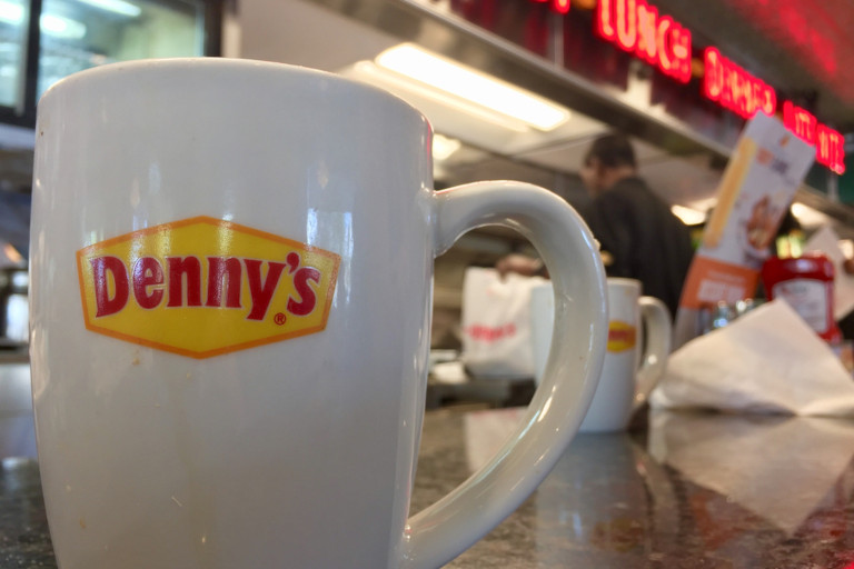 Cup with Denny's company logo sitting on a counter at the restaurant while a worker picks up a plate to deliver an order to a table in the background.