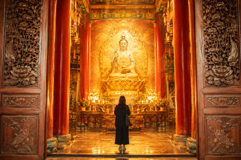 AI-generated image of a woman standing reverently before a brightly illuminated Guanyin statue inside an ornate Buddhist temple.