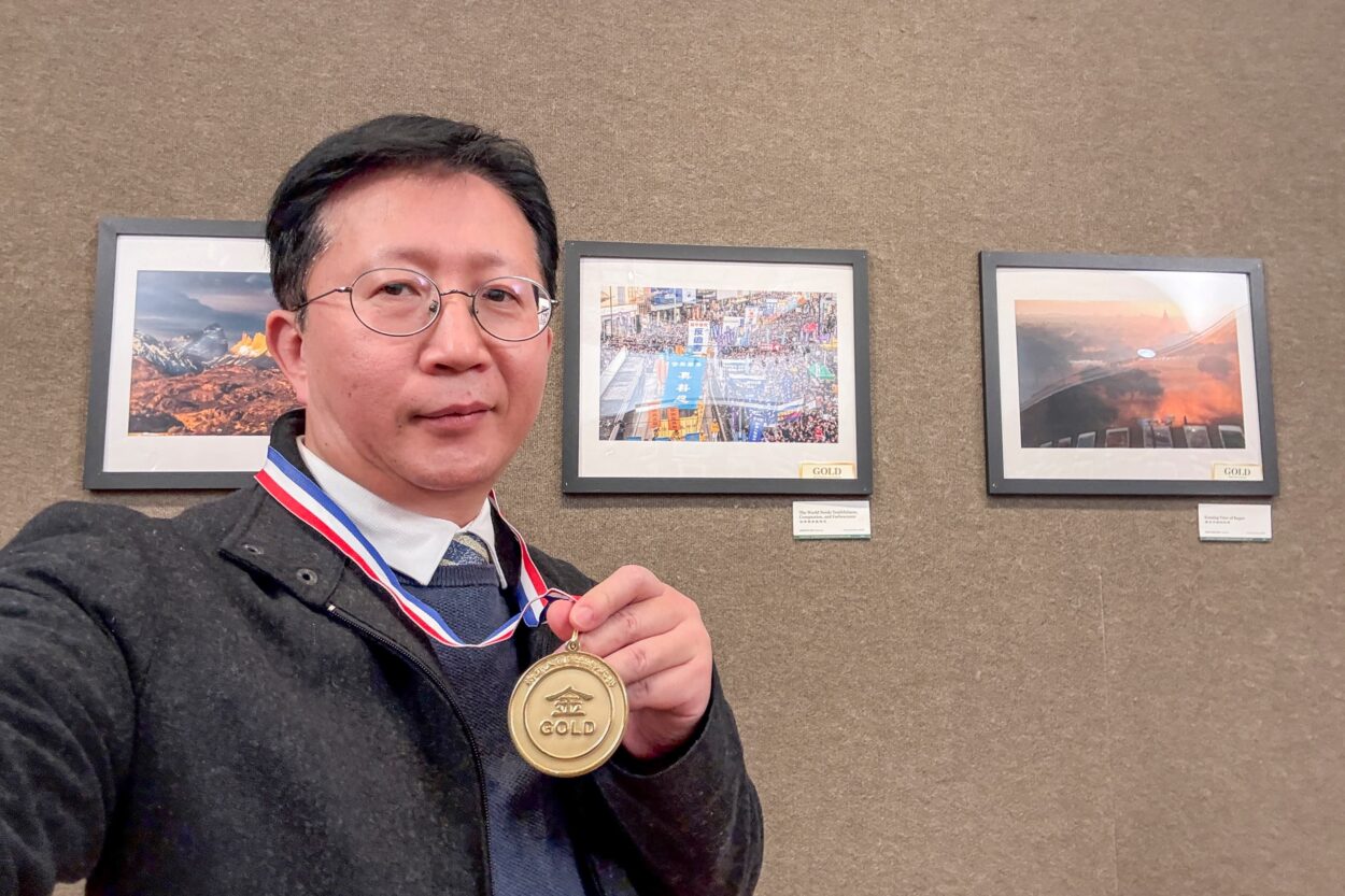 Hong Kong photojournalist, Adrien Yu holding up a price medal he won for a photography competition. Behind him his award winning photo in a frame.