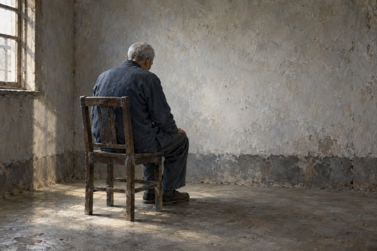 AI-generated image of an elderly Chinese man sitting alone on a wooden chair, facing the wall of a sparsely furnished room.