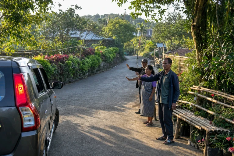 Mawlynnong residents calmly direct arriving cars and small taxis away from the village entrance while a quiet swept lane continues past flowering hedges and bamboo benches.