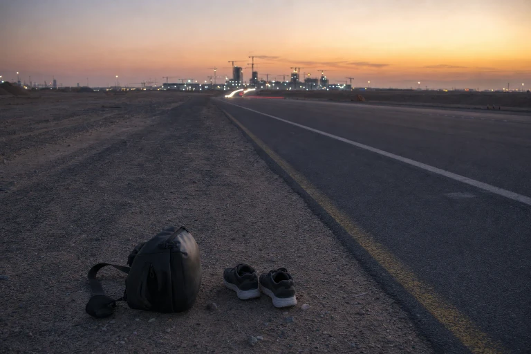An empty roadside at dusk holds a camera bag and a pair of walking shoes as a road leads toward distant construction lights.