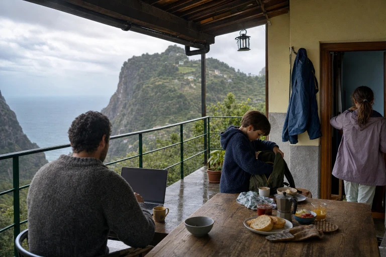 A family shares breakfast on a modest Madeira terrace while one parent works on an unreadable laptop and the children prepare for the day beside a drying jacket.
