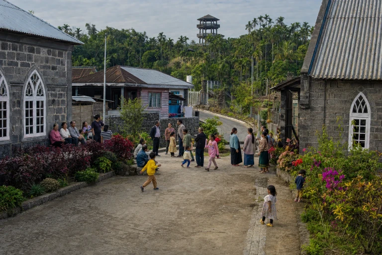 Families and neighbours share a quiet Sunday in an open space between stone churches and homes while children move among them, with a distant viewing tower and empty tourist approach beyond.