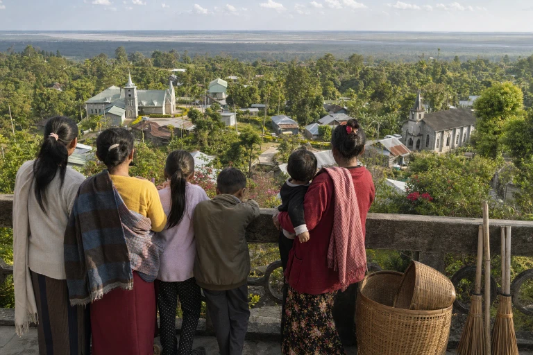 Children and adults gather on a village viewing tower and look across Mawlynnong’s homes, gardens, and quiet lanes toward the Bangladesh plains.